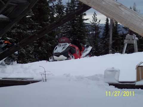 2004 Arctic Cat King Cat 900 - A snowmobile sits in a snowy landscape, partially obscured by snow and wooden structures.