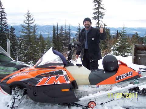2004 Arctic Cat King Cat 900 - Two snowmobiles are parked in a snowy outdoor setting, with a person standing behind them and trees in the background.