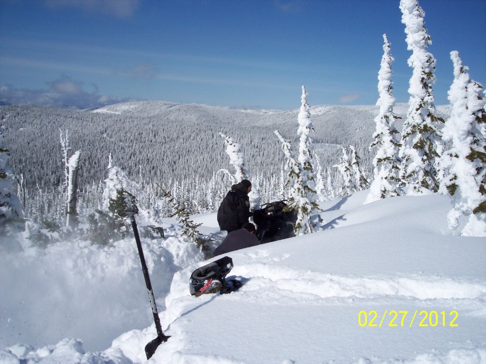 Snowy mountain scene with a person sitting in the snow next to a backpack and boots. A shovel is stuck in the snow