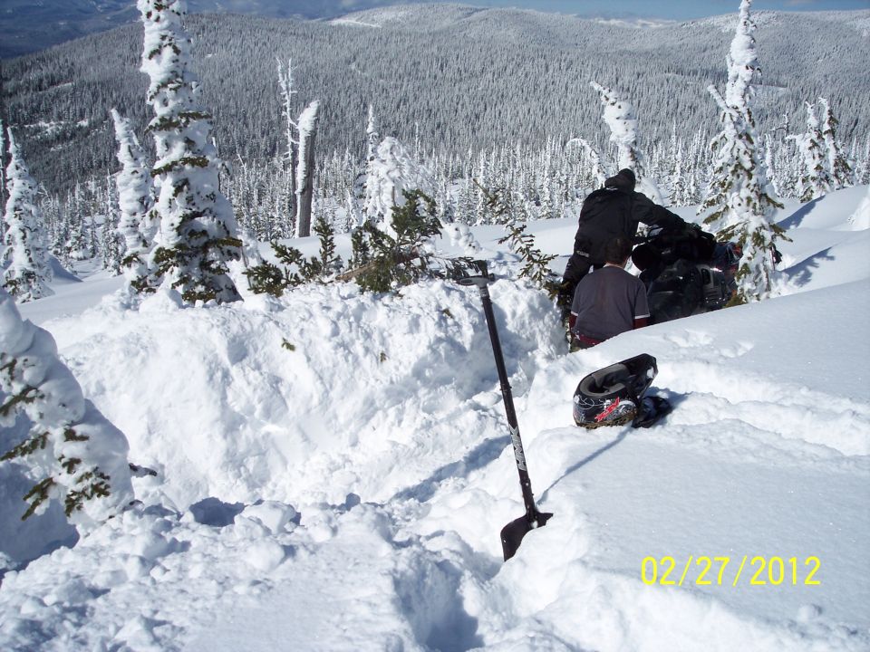 Snowmobile buried in snow, being dug out by two people with a shovel. Helmet in foreground.