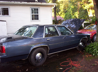 1991 Ford Crown Victoria LTD - A dark blue sedan is parked in a driveway next to a house, with another vehicle visible behind it.