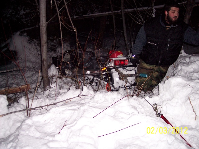 Snowmobile stuck in snow at night, being pulled by a winch with a person nearby.