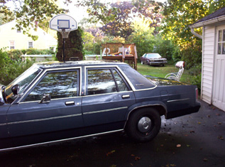 1991 Ford Crown Victoria LTD - A dark blue 1980s Ford Crown Victoria sedan is parked in a driveway next to a garage, with another car visible in the background.