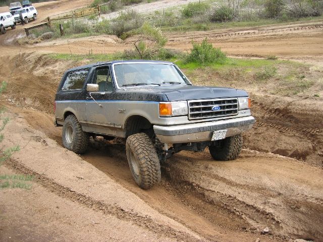 Gray-and-black lifted full-size Ford Bronco on a dirt off-road course, front three-quarter view