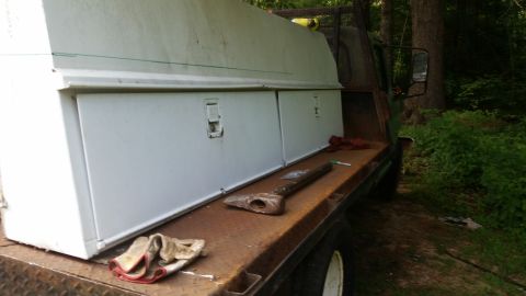 1978 Ford Bronco - A flatbed truck bed is shown with white storage boxes and a large axe resting on the rusty metal surface.