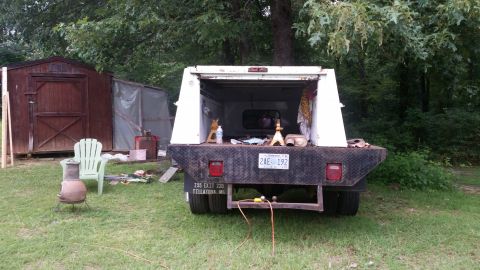 1978 Ford Bronco - A flatbed truck with a camper shell is parked in a grassy yard with sheds in the background.
