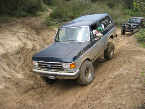 1989 Ford Bronco - A dark blue and silver Ford Bronco navigates a challenging dirt trail, with another vehicle visible behind it.