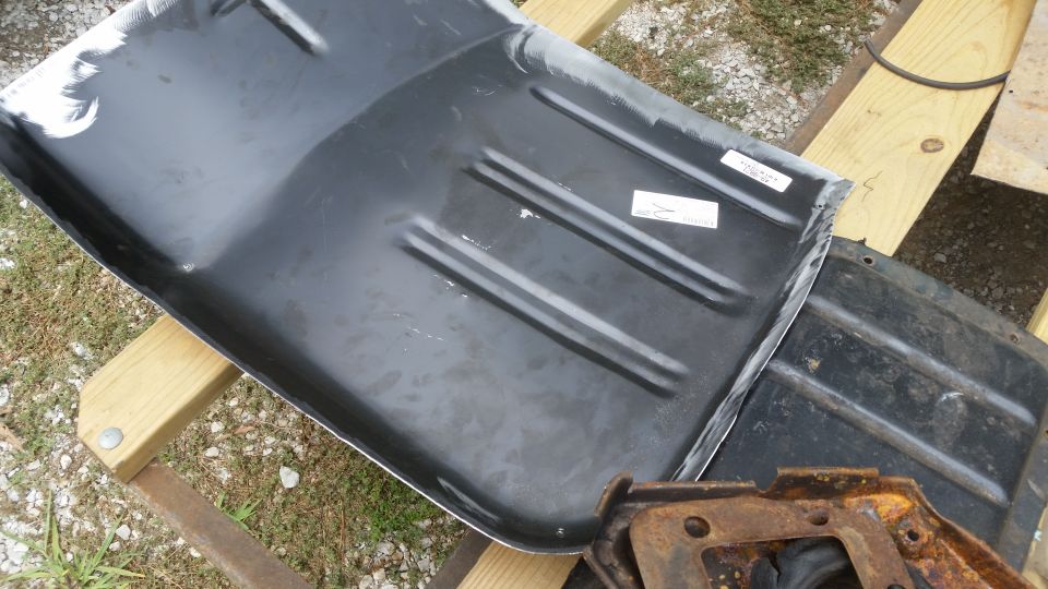 Close-up of a black metal vehicle floor pan with raised ribs, resting on wooden beams. Two white stickers are visible on the surface