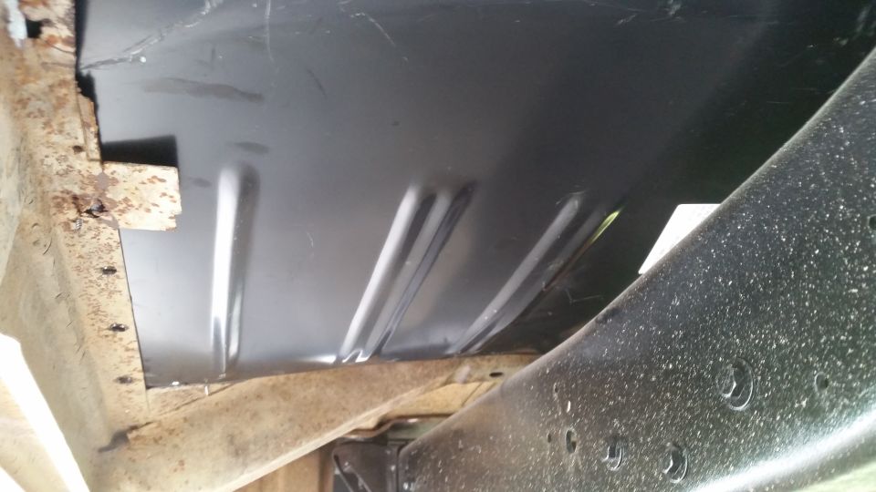 Undercarriage of a vehicle showing a black metal floor panel with ribbed reinforcement and rusty frame components