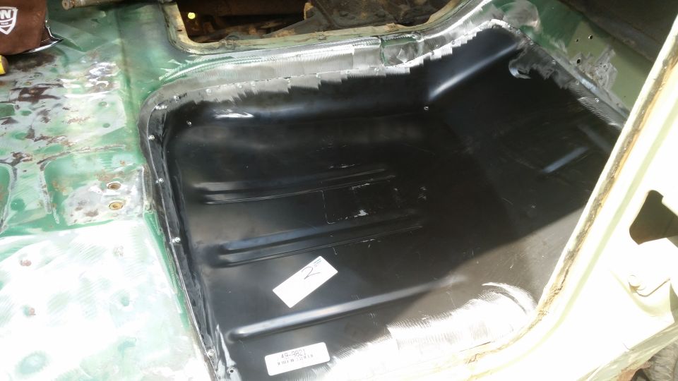 Interior floor pan of a vehicle undergoing repair, showing new black metal paneling and green primer