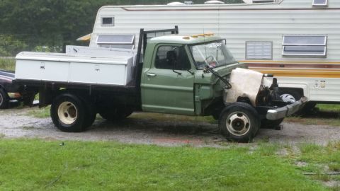 Green flatbed truck with a white toolbox on the bed, parked next to a camper. Front end is partially disassemb