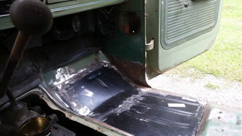 Green vehicle interior floor with exposed metal and rust patches, showing a floor shifter and open door