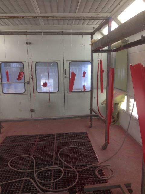 1964 Ford Fairlane - A spray booth is set up with various red parts suspended for painting, with a metal grate floor and a coiled hose.