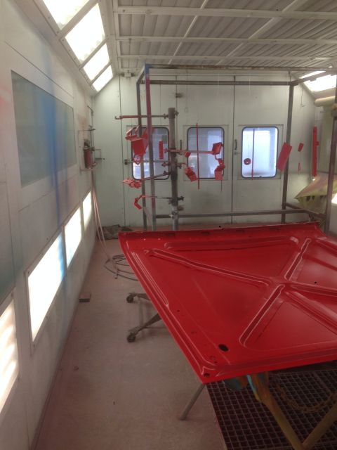 1964 Ford Fairlane - A freshly painted red car hood rests on a stand inside a brightly lit automotive paint booth.
