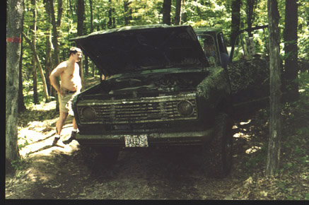 1991 Ford Bronco - Off-road truck with hood open, covered in mud and foliage, parked in a wooded area.
