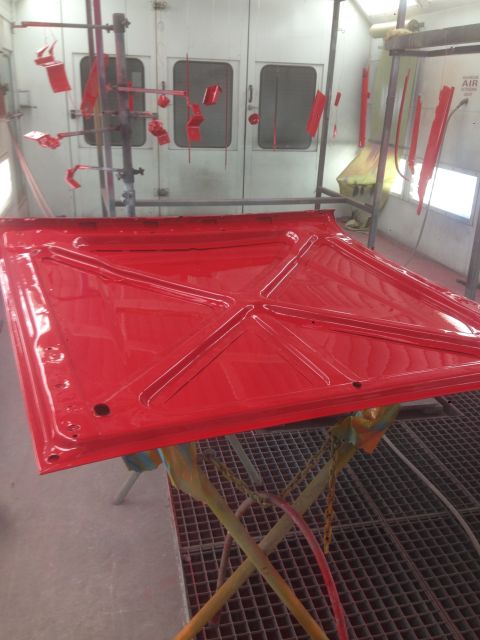 1964 Ford Fairlane - A freshly painted red car hood rests on a stand inside a professional paint booth, awaiting reassembly.