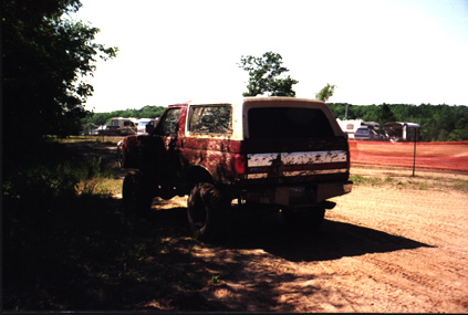 1991 Ford Bronco - Muddy, lifted 1991 Ford Bronco seen from the rear on a dirt track with other vehicles in the background.