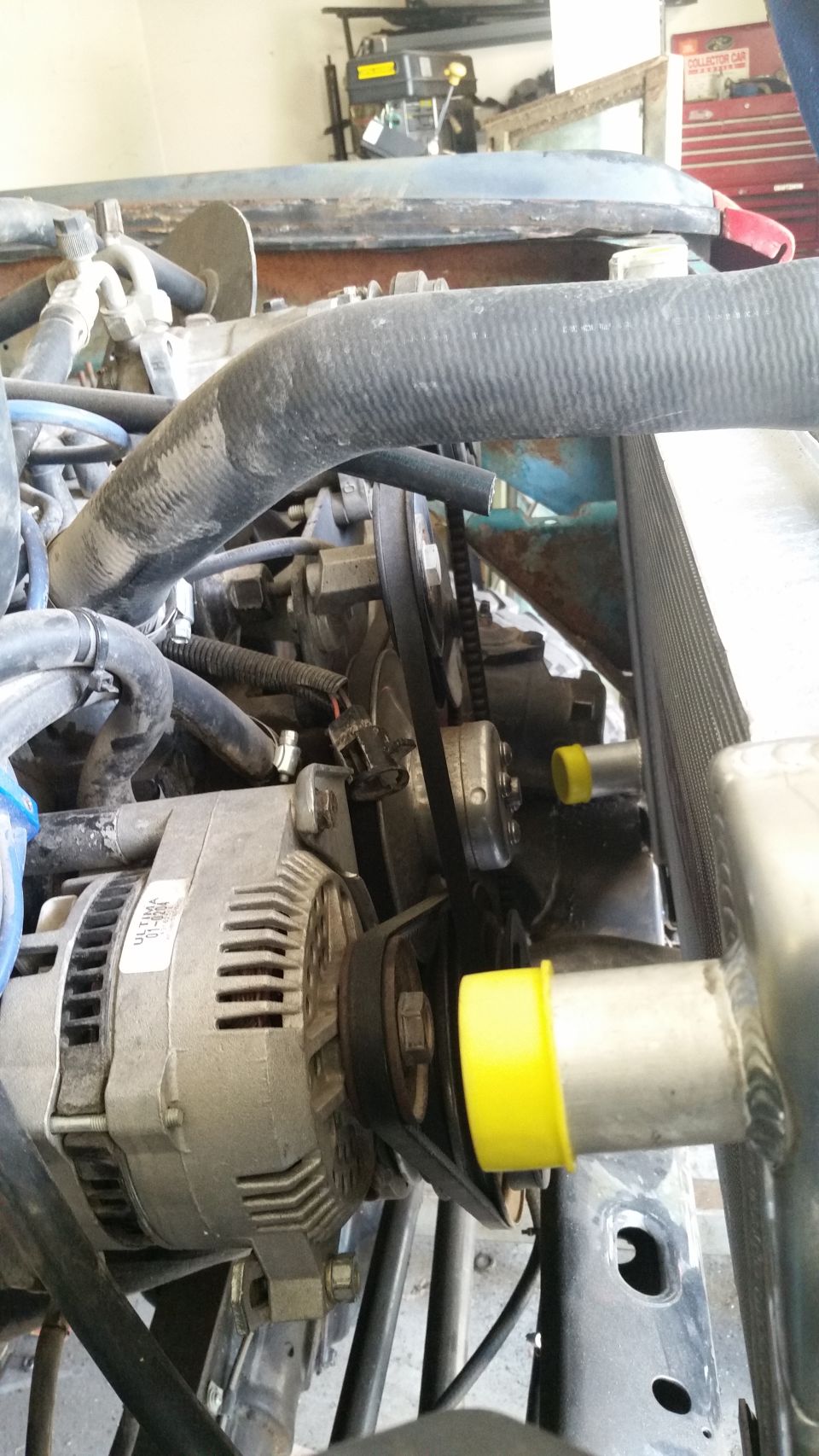 Engine bay showing alternator, belts, radiator hoses, and part of the radiator. Yellow caps on hoses