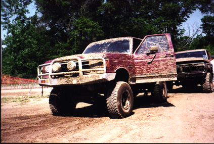 1991 Ford Bronco - A muddy 1991 Ford Bronco sits with its door ajar, parked beside another truck on a dirt track.