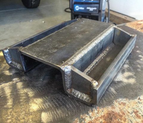 1979 Ford Bronco - A custom-fabricated steel part with prominent welds sits on a textured surface, with a welding machine in the background.