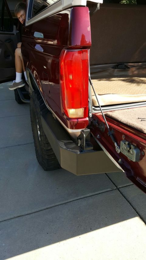 1996 Ford Bronco - A young person sits in the open door of a red pickup truck, with the rear bumper and taillight in view.