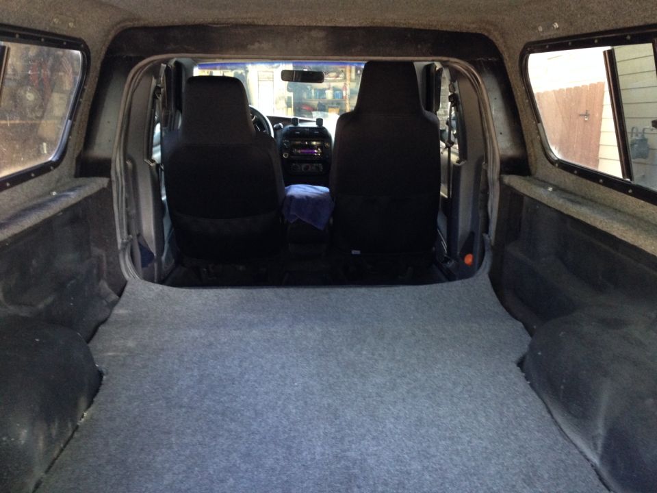Interior view of a pickup truck bed with carpet, showing the back of the front seats and a camper shell