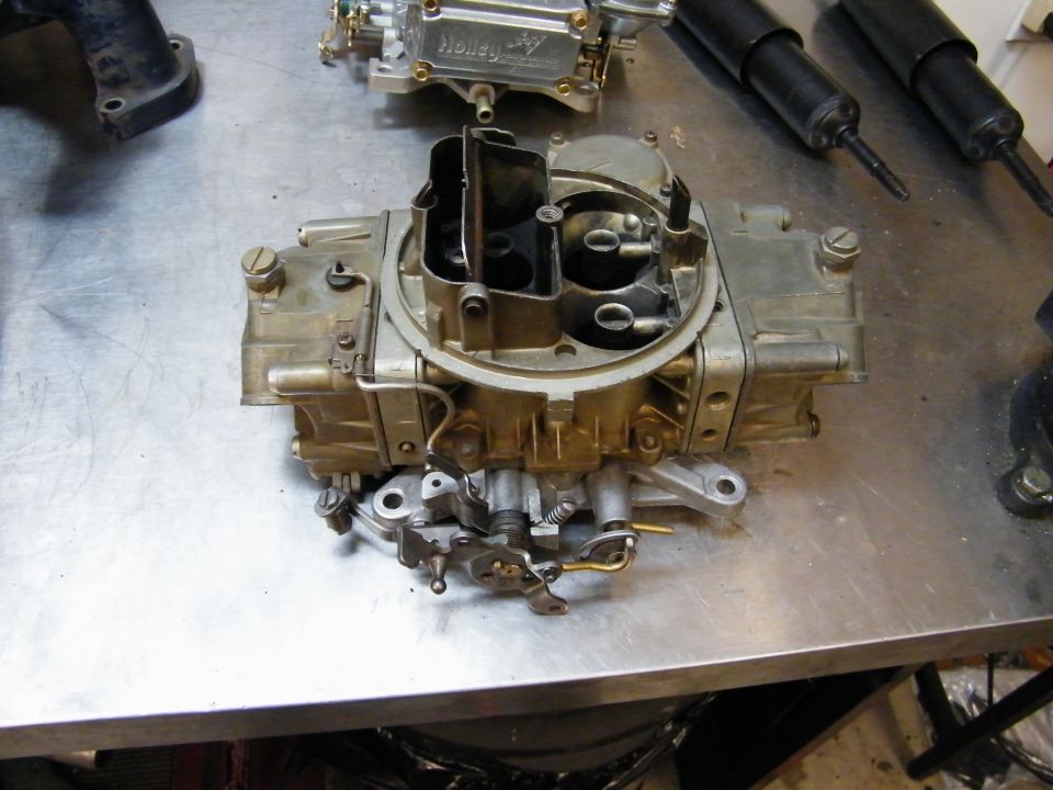 Holley carburetor on a metal workbench with another carburetor and two black shock absorbers in the background