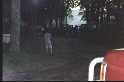 1991 Ford Bronco - A group gathers around vehicles in a wooded area, with a red vehicle part visible in the foreground.