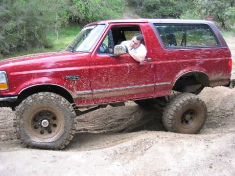 1989 Ford Bronco - A red Ford Bronco navigates a muddy off-road trail, its tires caked in dirt.