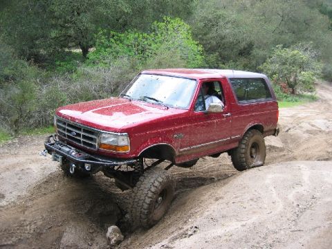 1989 Ford Bronco - A red Ford Bronco navigates a challenging dirt trail, its front grille and headlights visible.