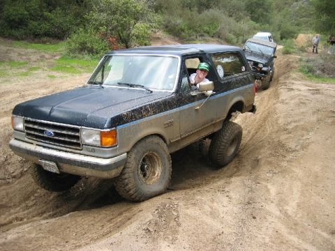 1989 Ford Bronco - Two SUVs off-roading on a dirt path, with the lead Bronco in the foreground.