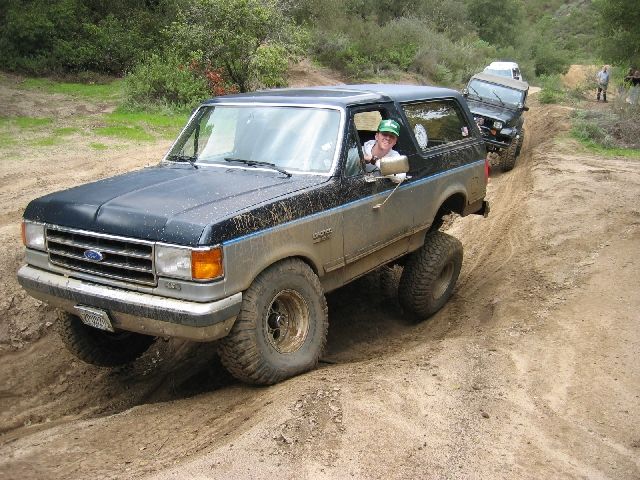Lifted dark blue Ford Bronco with top removed flexing over a rutted dirt trail