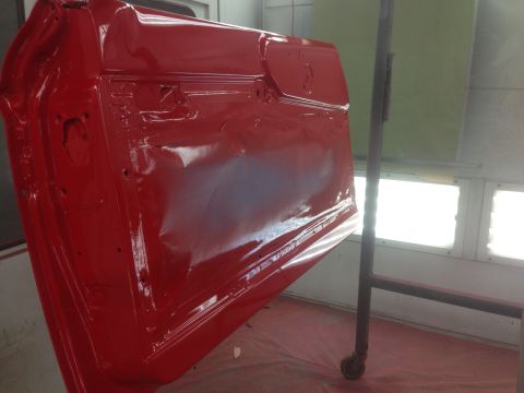 1964 Ford Fairlane - A red car door is being painted in a booth, suspended by a metal rack.