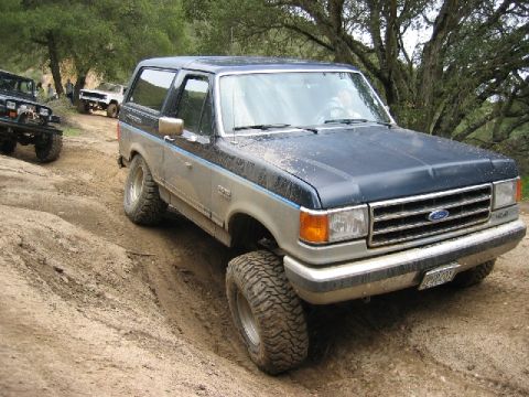 1989 Ford Bronco - A lifted Ford Bronco navigates a muddy off-road path, with other vehicles following.