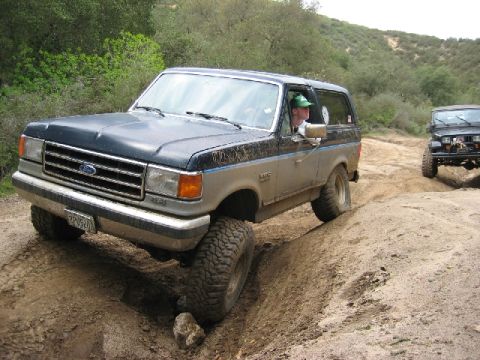 1989 Ford Bronco - Two off-road vehicles, a Ford Bronco and a Jeep, navigate a dusty trail on a hillside.