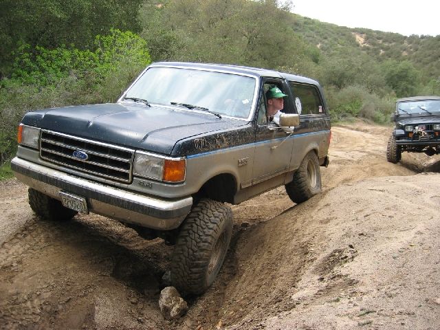 Blue and tan Ford Bronco flexing over ruts on a dirt trail with a Jeep behind