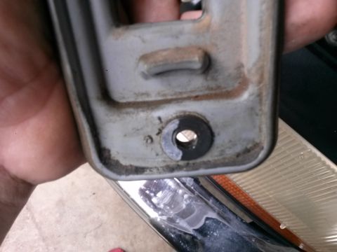 1996 Ford Bronco - A hand holds a metal bracket, showing a bolt hole and some wear. A vehicle's headlight assembly is partially visible in the background.