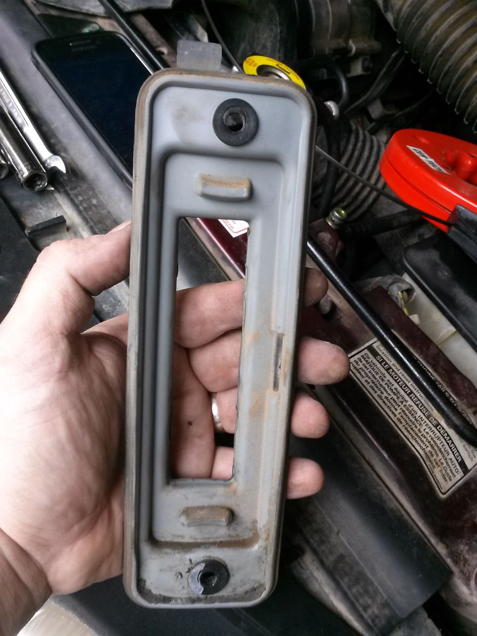 Hand holding a gray metal bracket with visible rust and a black rubber grommet, in a vehicle engine bay