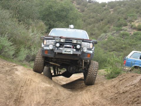 1989 Ford Bronco - This lifted white SUV navigates a challenging dirt trail, equipped with numerous off-road lights and a front-mounted winch. A second blue S…