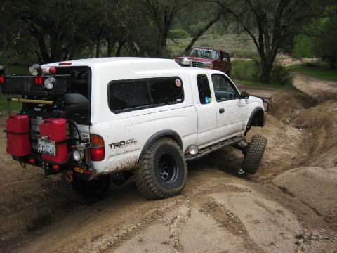 1989 Ford Bronco - Modified white Toyota Tacoma navigating an off-road trail with a red SUV in the background.
