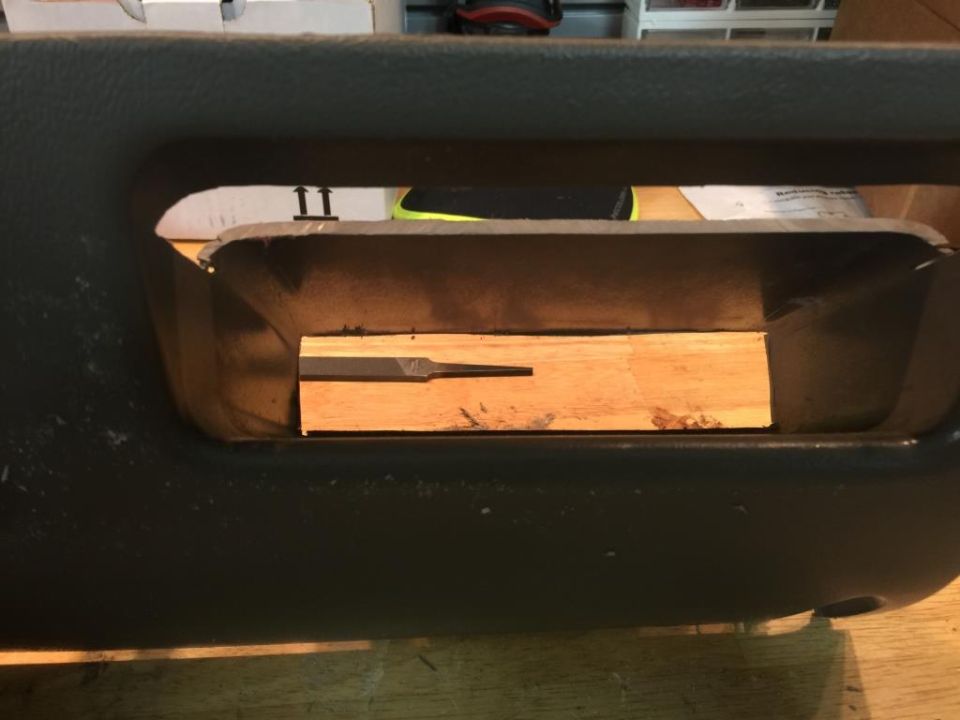 Dark gray interior panel with a rectangular opening containing a wooden block and a file