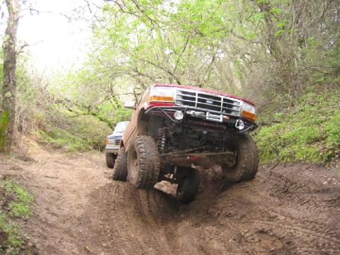 1989 Ford Bronco - Two trucks navigate a challenging muddy trail surrounded by trees.
