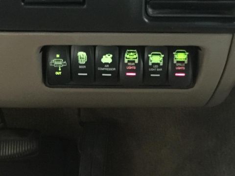 1996 Ford Bronco - A row of aftermarket rocker switches with illuminated labels and icons are installed in a vehicle's dashboard.