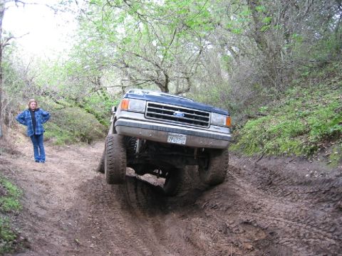 1989 Ford Bronco - A dark blue Ford Bronco navigates a challenging muddy off-road trail with its front passenger wheel lifted.