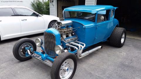 1930 Ford Coupe - A bright blue 1930 Ford Coupe hot rod is parked in front of a garage, with a white modern sedan beside it.