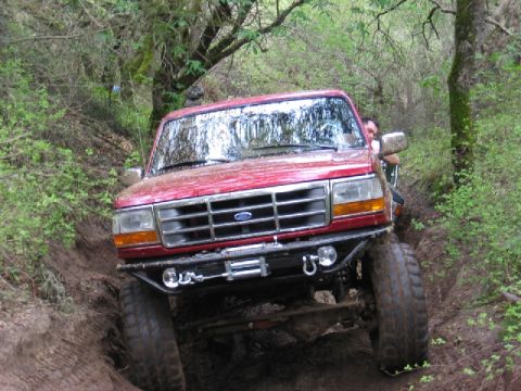 1989 Ford Bronco - Red 1989 Ford Bronco navigating a difficult off-road path with mud and trees.