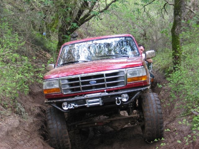 Muddy red lifted Ford 4x4 with winch bumper climbing a rutted wooded trail