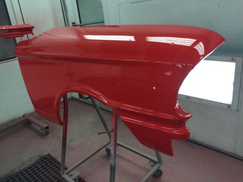 1964 Ford Fairlane - A freshly painted red fender, likely from a 1964 Ford Fairlane, is mounted on a stand in a spray booth.