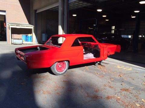 1964 Ford Fairlane - A red car body, stripped down and on wheels, sits outside a garage, likely undergoing restoration.