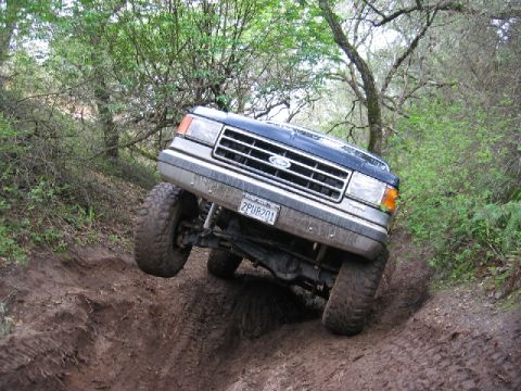 1989 Ford Bronco - This 1989 Ford Bronco navigates a challenging, muddy off-road path surrounded by trees.